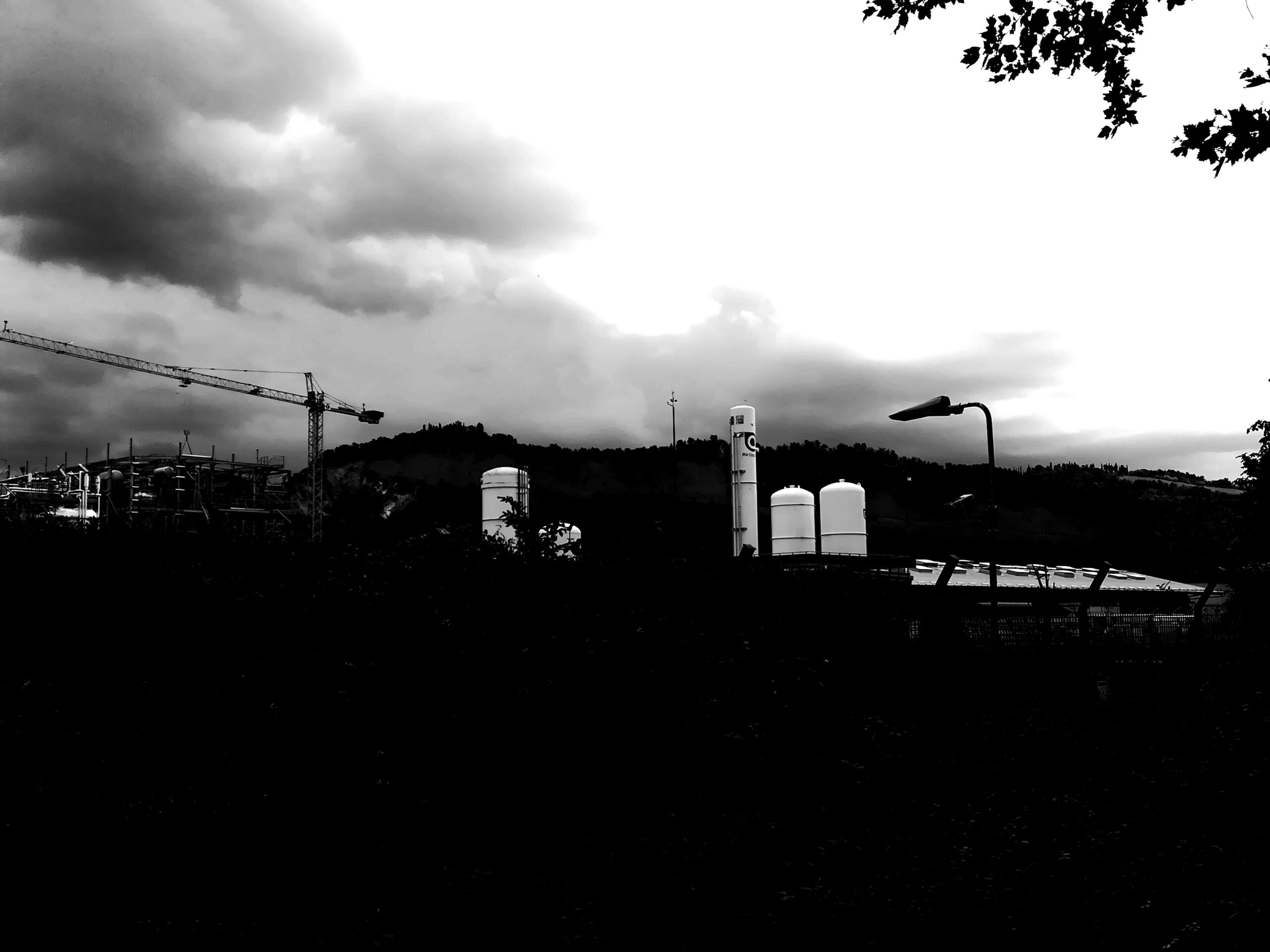 Fotografia in bianco e nero ad alto contrasto di un paesaggio industriale cupo sotto un cielo tempestoso. In primo piano domina un'ombra nera e fitta che nasconde il terreno; sullo sfondo emergono silos bianchi, una gru da cantiere e un lampione isolato contro il profilo scuro delle colline.
