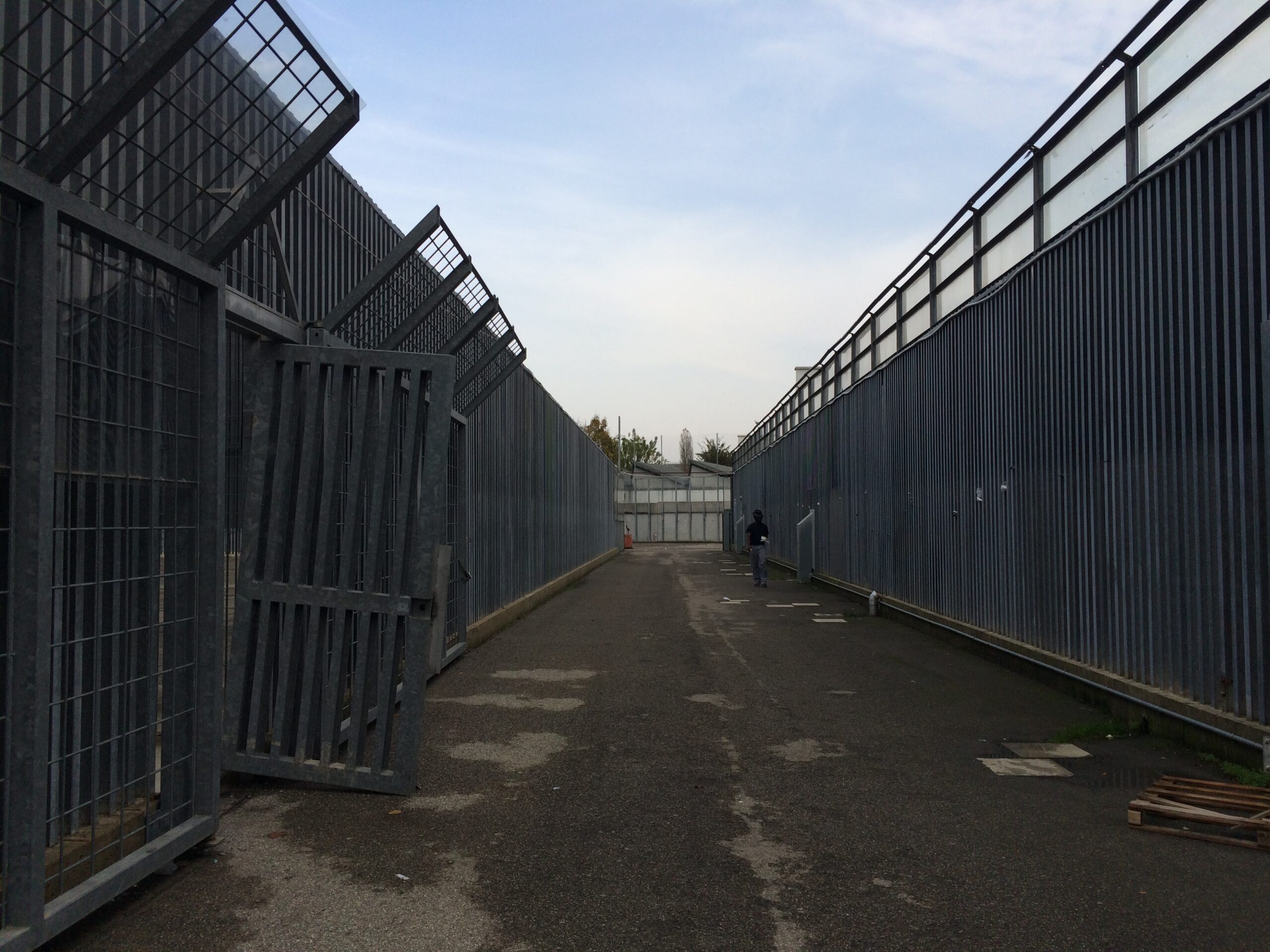 Biopolitica, emergenza e mobilità nell'ex CIE di Bologna: un lungo e desolato corridoio in asfalto, delimitato da altissime recinzioni metalliche e sbarre di ferro con griglie anti-scavalco.